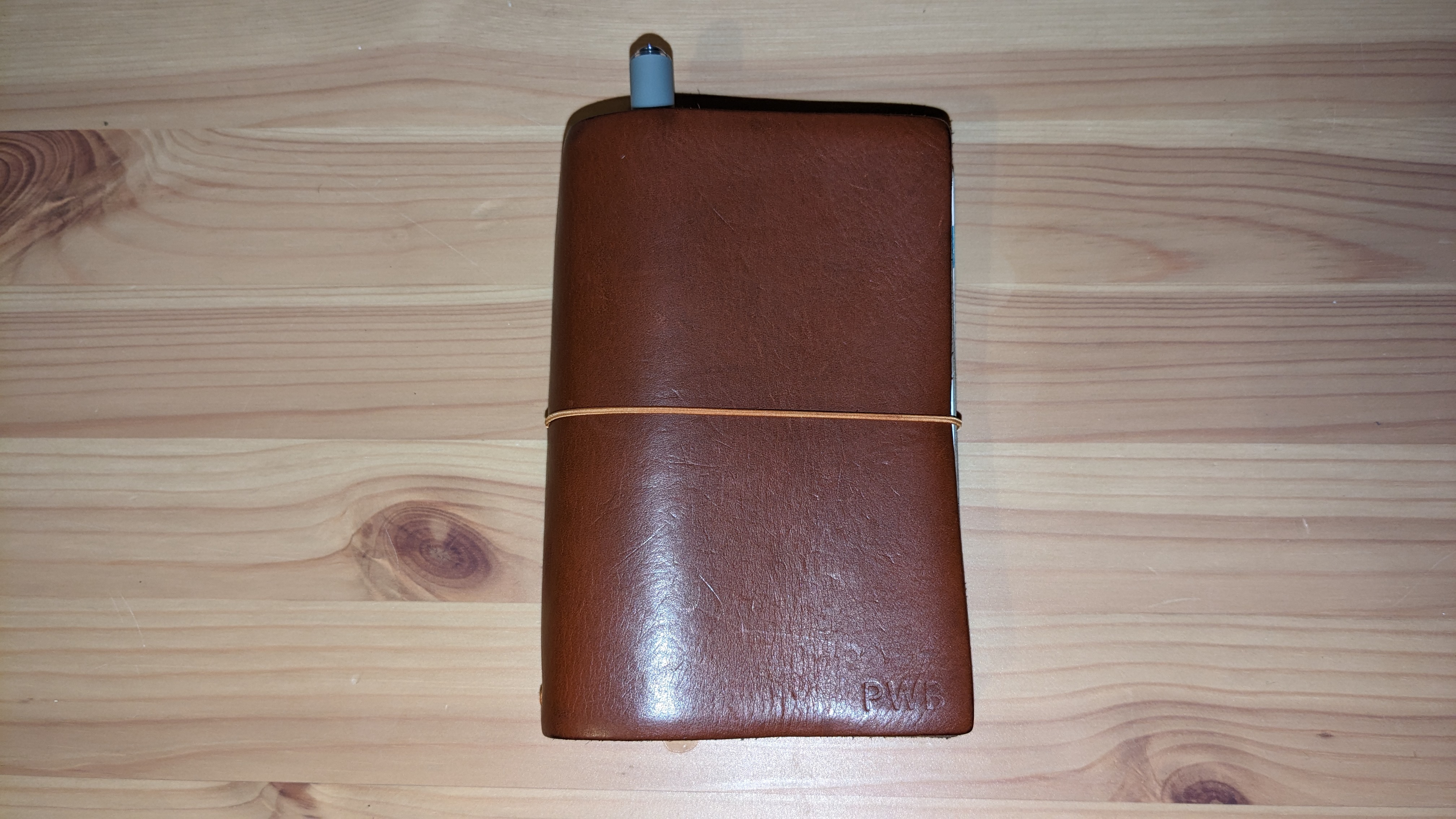 A notebook on a wooden table. The cover is made of scuffed brown leather, with an orange elastic band, and the initials 'PWB' are faintly visible in the corner.